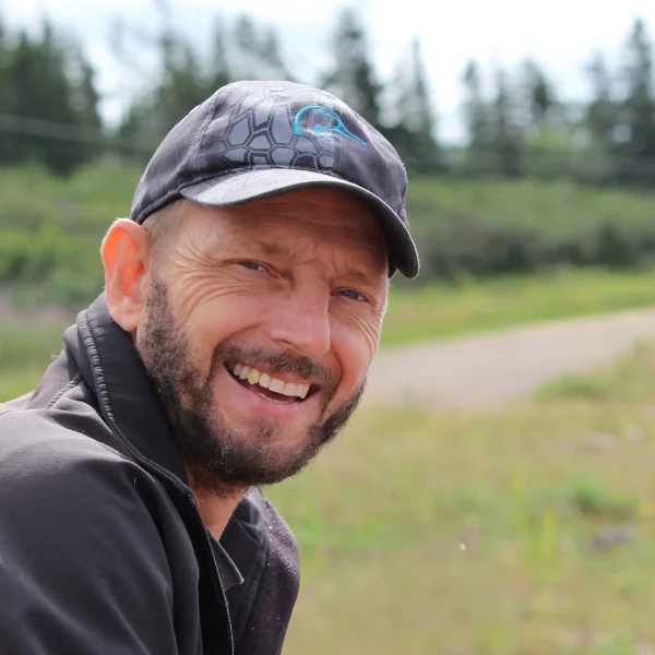 Ken, owner of Maritime Sharpening in Halifax, smiling at the camera. Wearing a black cap and black jacket. In the background some trees and grass.